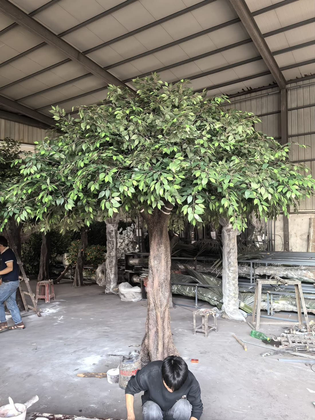 Large-canopy artificial ficus tree being checked at full height by a worker in the Everflorae workshop, with the trunk in bolt-together segments